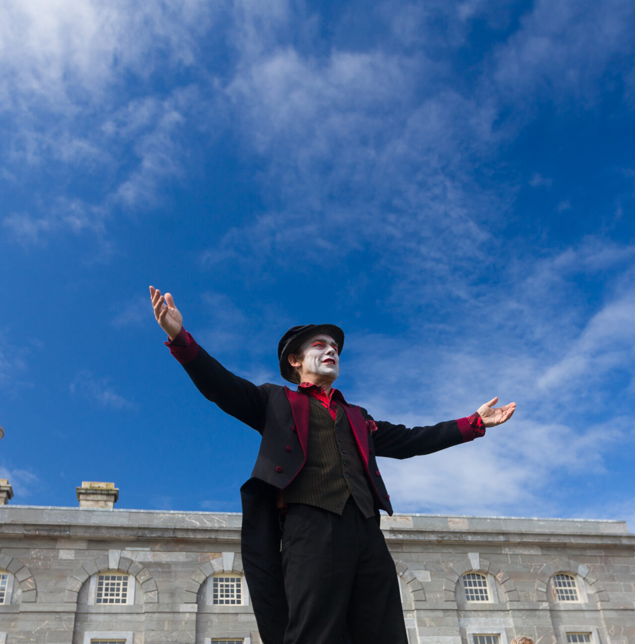 Invisible Circus Ringleader, Doug Francis, stands tall with open arms and a smiling face, wearing a tail suit and bowler hat, with traditional clown makeup, set against a backdrop of blue sky and grey buildings.